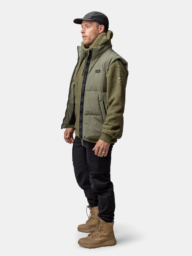 Man i grön Halti Penger Classic Puffer Väst Herr, fleecejacka, svarta byxor, solbruna stövlar och svart keps.