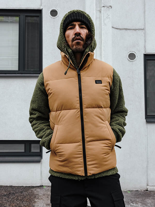 En man i brun Halti Penger Classic Puffer Väst Herr står utanför en vit byggnad med fönster.