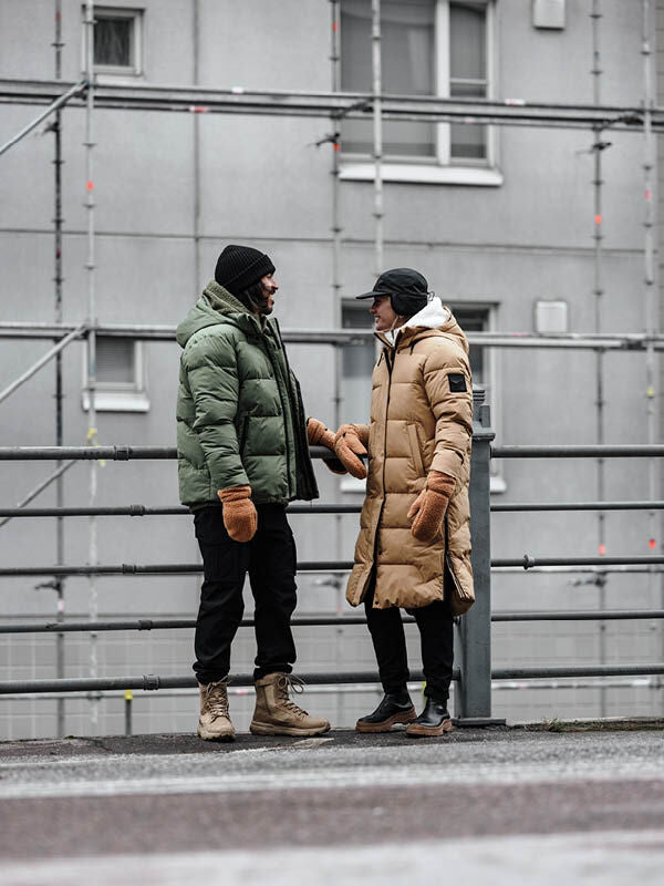 Två personer i Halti Brahe Puffer Long Vinterjacka Dam pratar på en trottoar med byggnadsställningar bakom sig.