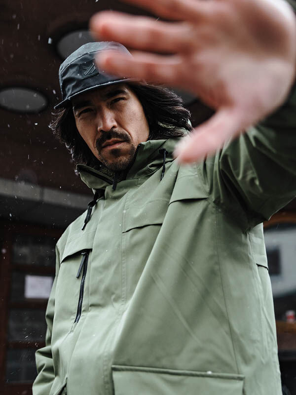 Man i grön Halti Tokoi II DX herrparkas och svart keps lyfter handen mot kameran, utomhus med allvarlig blick.