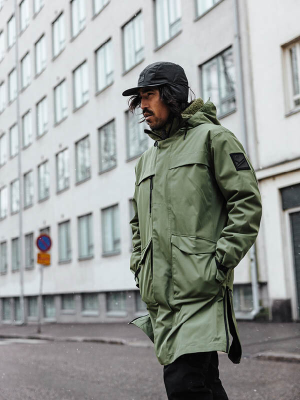 En person i grön Halti Tokoi II DX Parkajacka Herr och svart keps går förbi flerfamiljshus i staden.