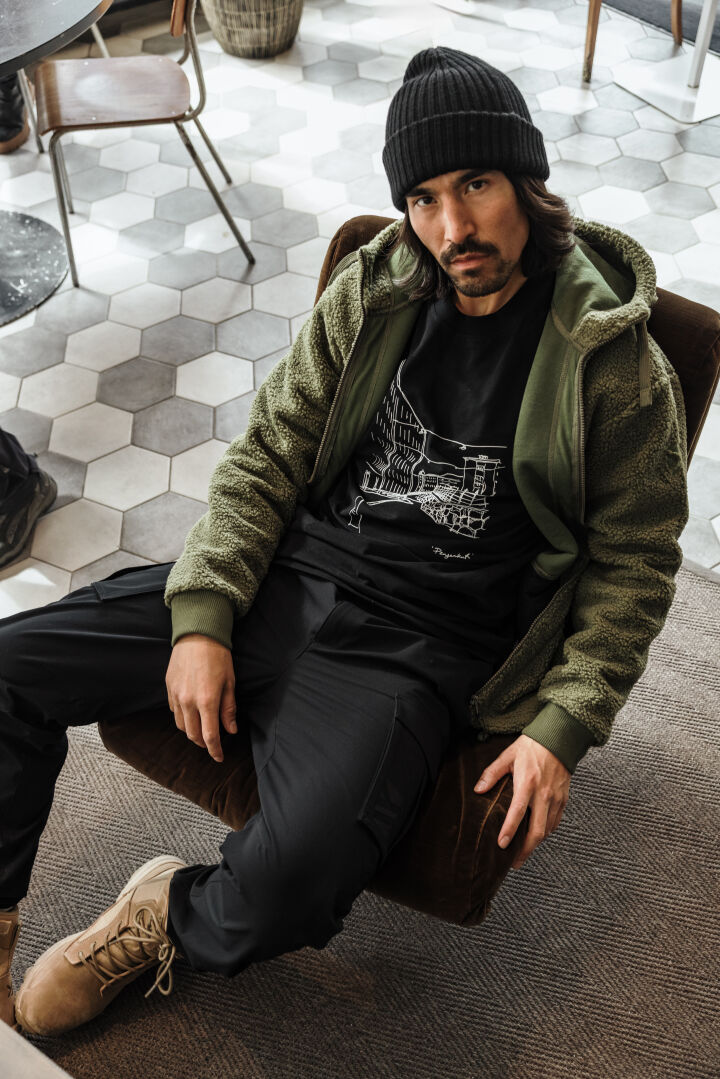 En man i grön Halti Lykky Fleecejacka Herr, svart mössa och stövlar sitter på en stol och tittar in i kameran.