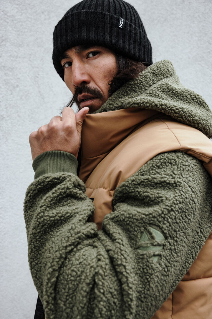 Man bär Halti Lykky Fleecejacka Herr i grönt och brunt och en svart mössa och håller i sin jackkrage.