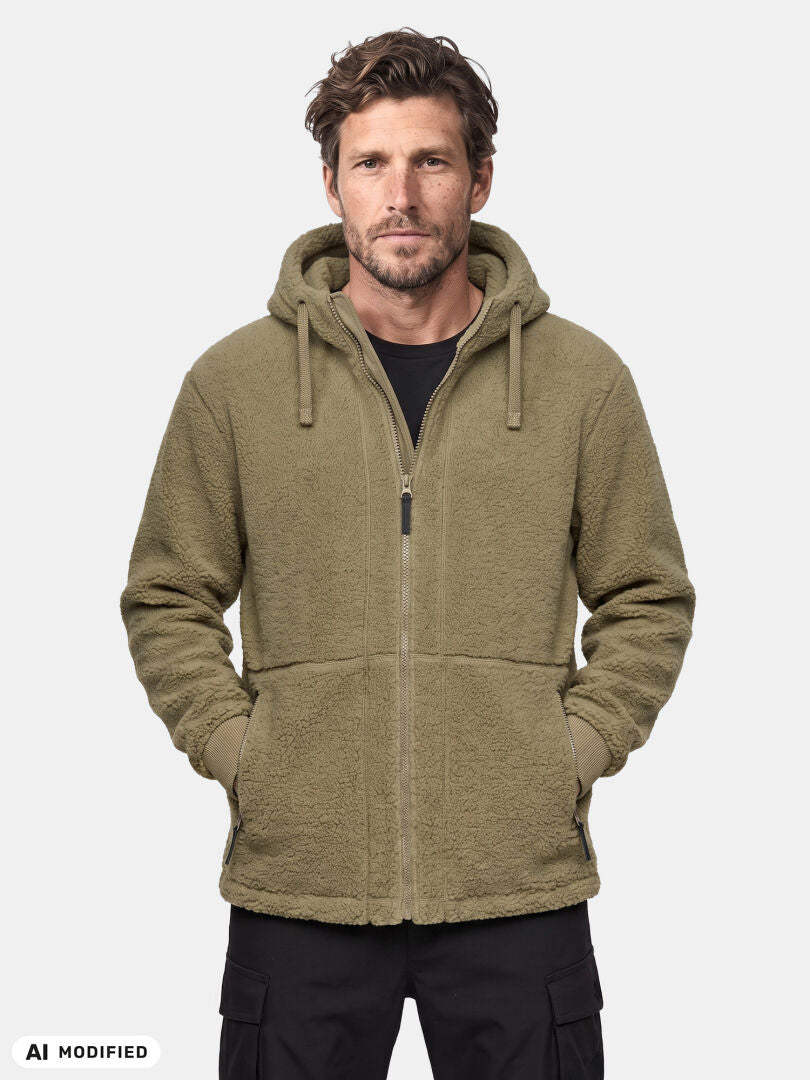En man bär en Halti Lykky Fleece Jacka Herr i olivgrönt, med händerna i fickorna, stående framför en enfärgad bakgrund.