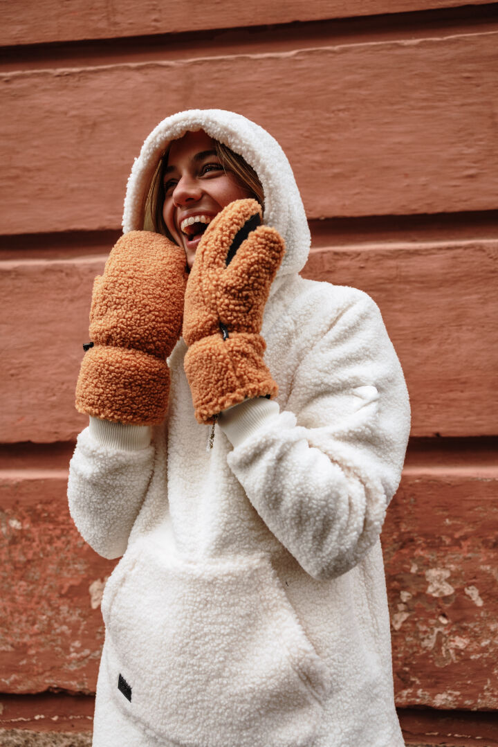 En kvinna i Halti Lykky Fleecetröjä Dam och bruna vantar ler framför en rödbrun strukturerad vägg.