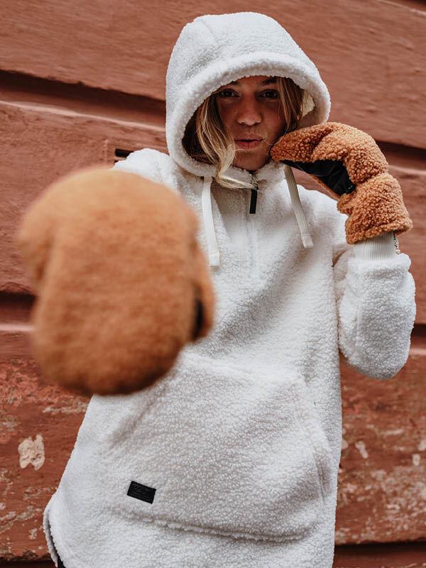 En kvinna i Halti Lykky Fleecetröjä Dam och bruna vantar poserar med uppsträckta nävar framför en röd texturerad vägg.