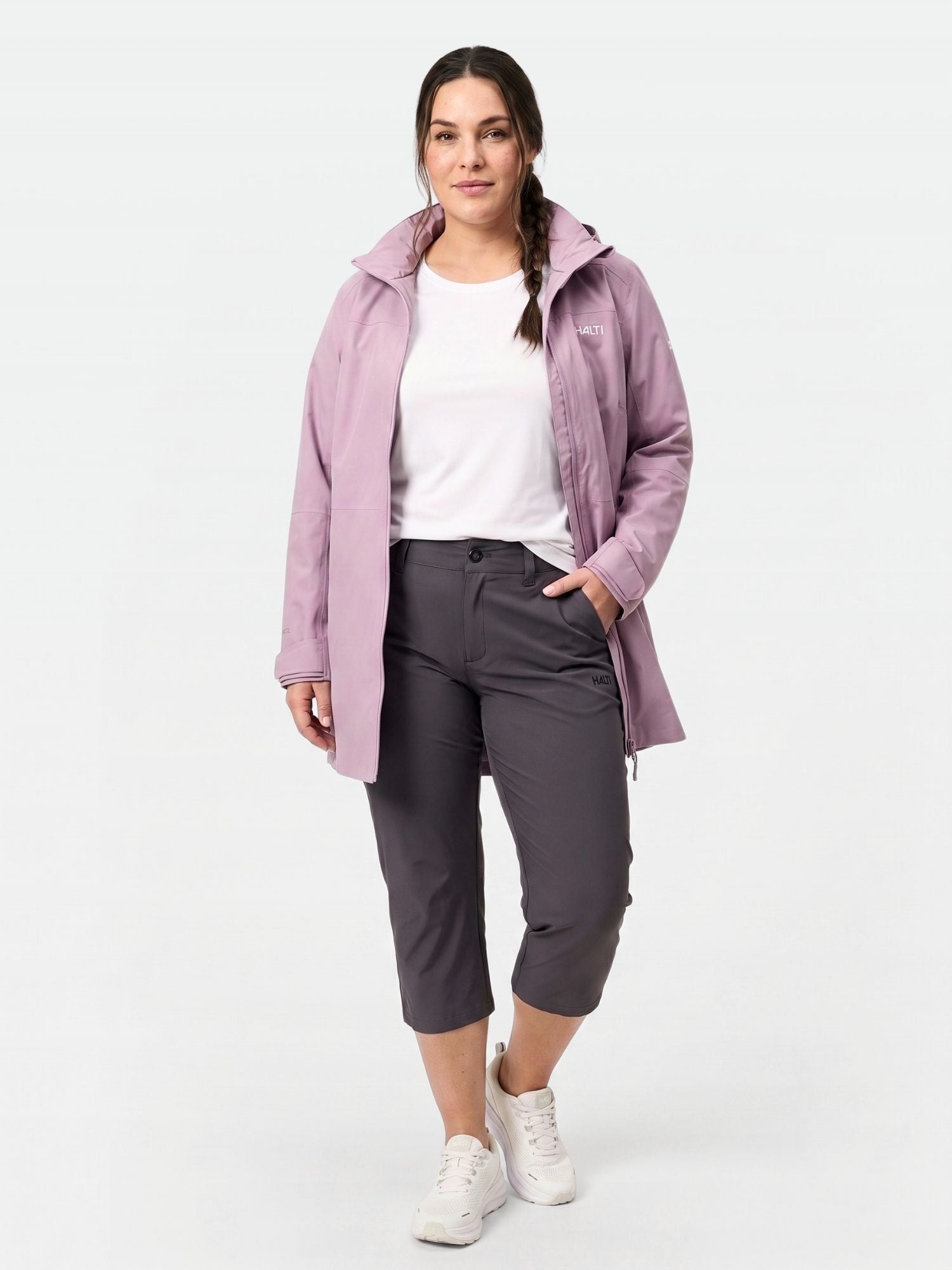 En kvinna vänder sig framåt i Halti Drive X-stretch Capribyxor Dam Plus med UPF 50+, ljuslila jacka och vita sneakers.