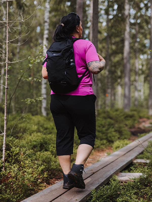 En person i rosa skjorta, svart ryggsäck och Halti Drive X-stretch Capribyxor Dam Plus går på en skogsstig av trä.