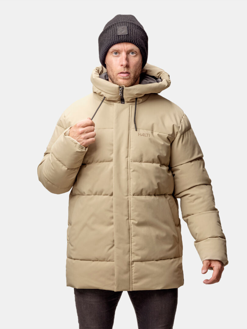 En man i beige Halti Sammal Vadderad Parkajacka och mörk stickad mössa står mot en ljus bakgrund.
