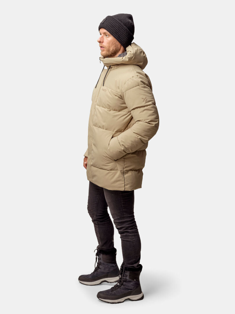 En man i brun Halti Sammal Vadderad Parkajacka, svart mössa, byxor och stövlar står med händerna i fickorna, vänd åt vänster.