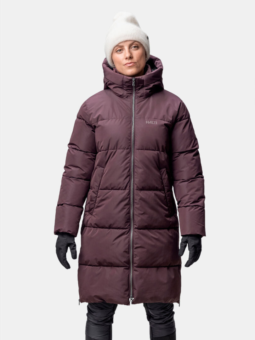 En person i en lång, vinröd Halti Sammal Vadderad Parkajacka Dam och vit mössa står vänd framåt.