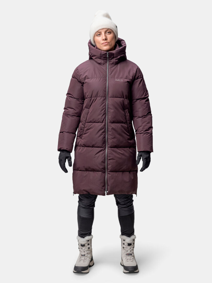 En person har på sig en rödbrun Halti Sammal Vadderad Parkajacka Dam, svarta handskar, vit mössa, svarta byxor och grå vinterkängor.