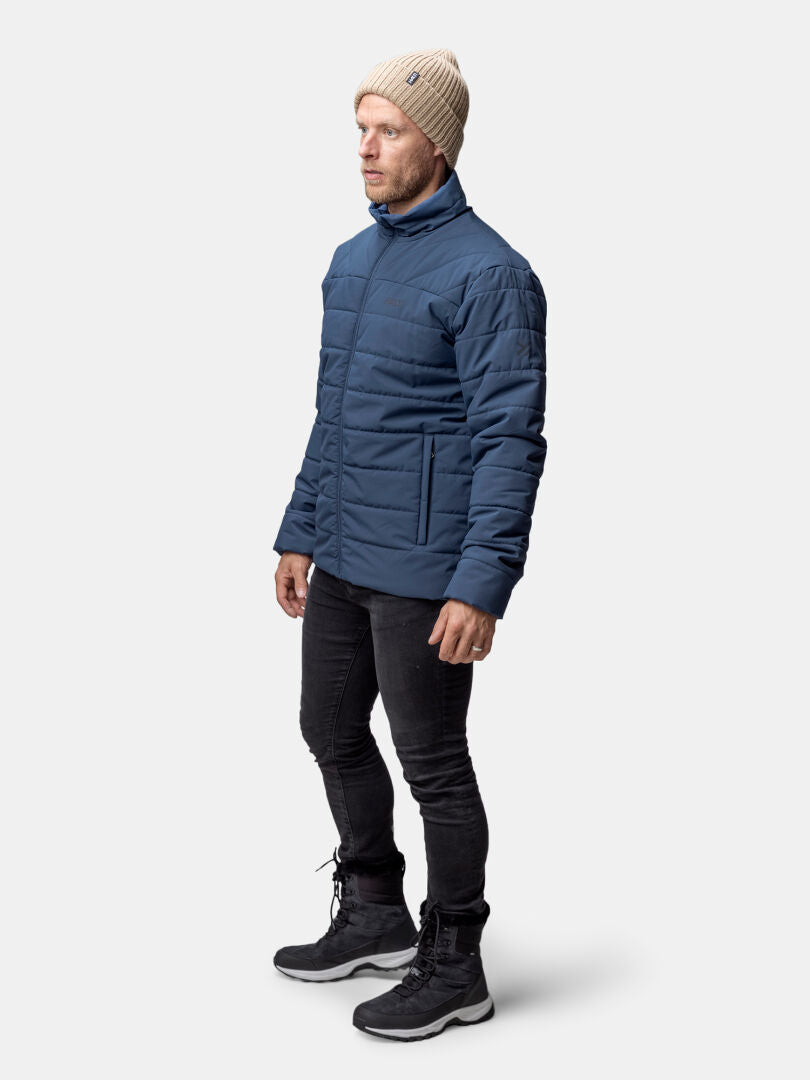Man i beige stickad mössa, Halti Haapa Vadderad Jacka Herr, svarta jeans och kängor står mot en enfärgad bakgrund.
