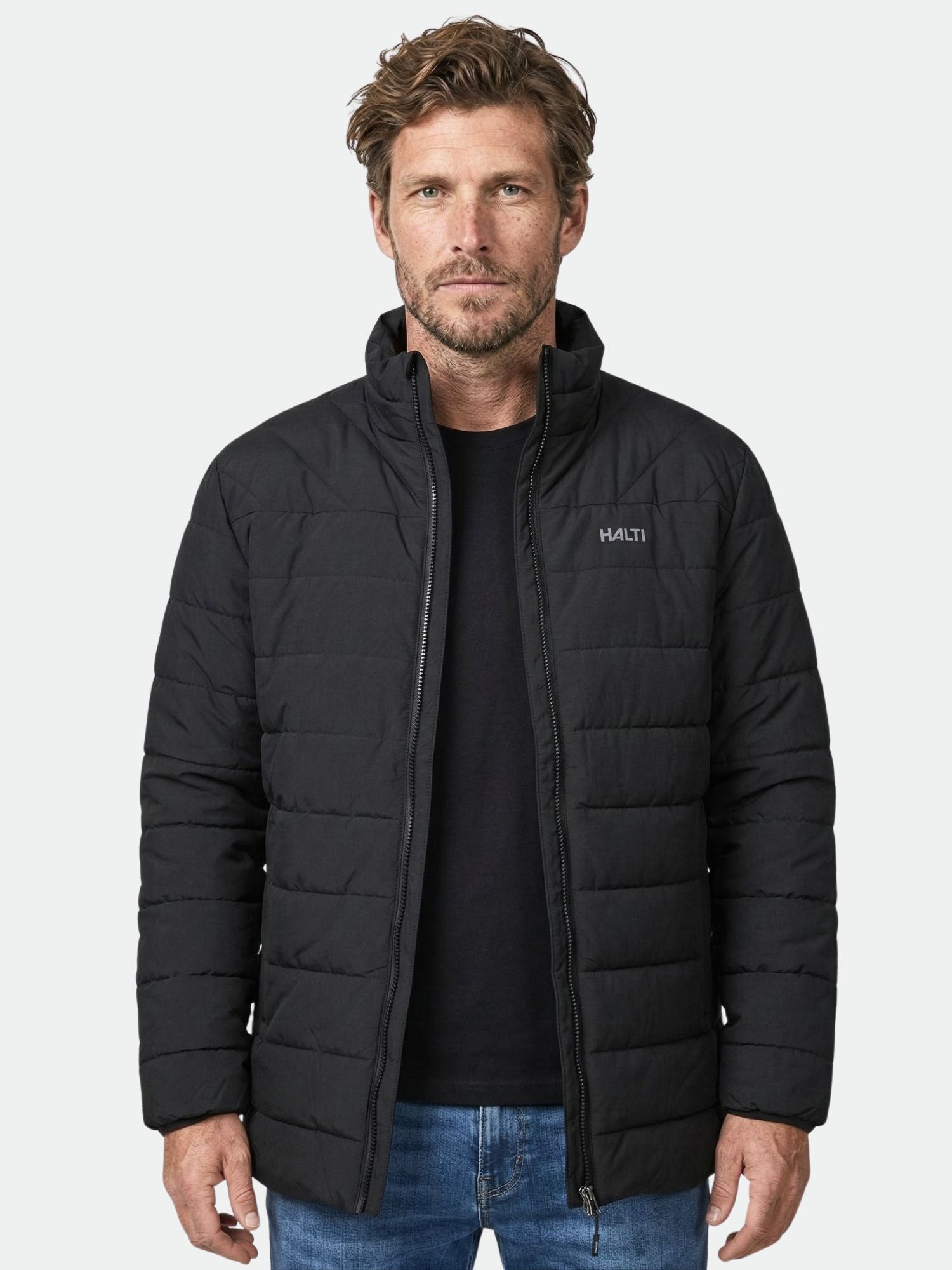 En man bär en Halti Haapa Vadderad Jacka Herr i svart och blå jeans, vänd framåt mot en ljus bakgrund.