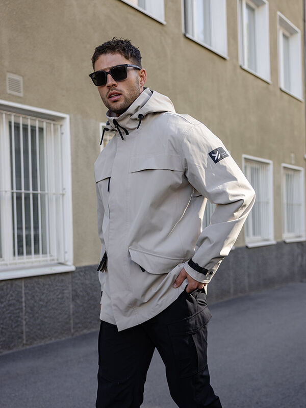 En man i Halti Tokoi DX Jacka Herr står på en trottoar vid en beige byggnad med gallerförsedda fönster.