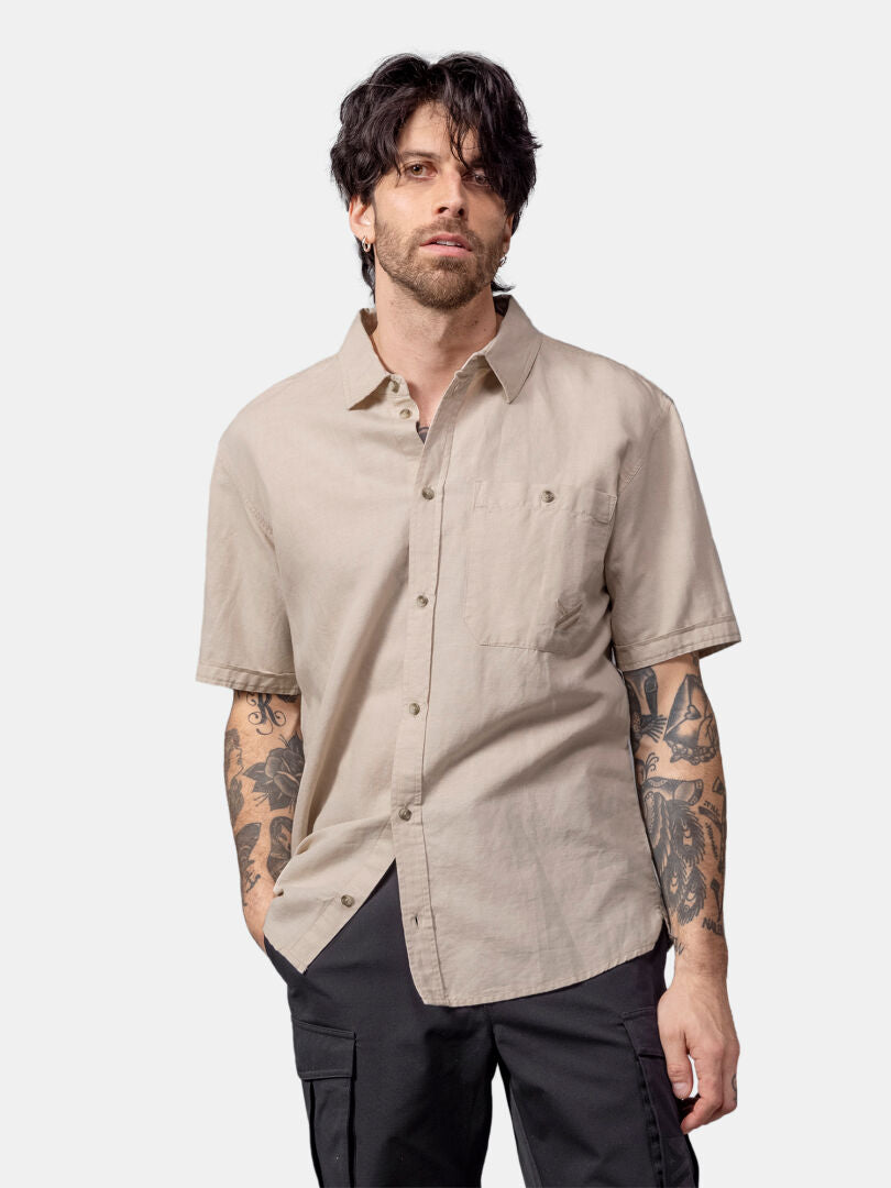 En man med mörkt hår och tatueringar bär en Halti Notsgi Skjorta Herr i beige och svarta byxor, stående mot en enfärgad bakgrund.