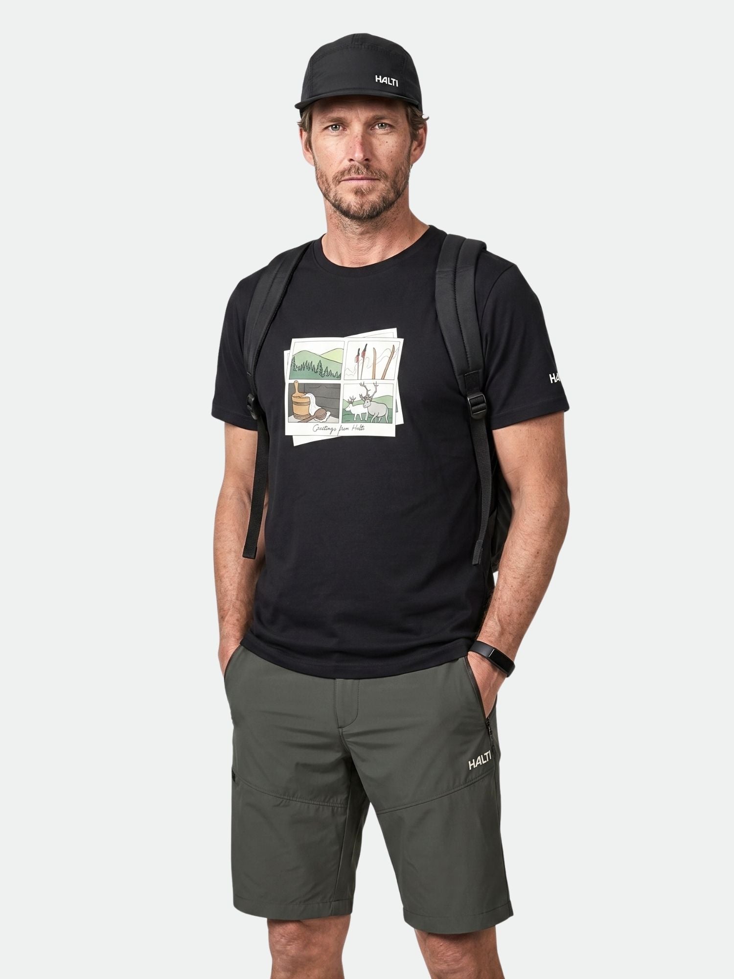 En man i svart keps, Halti Matka II T-skjorta Herr, gröna shorts och ryggsäck står med händerna i fickorna.