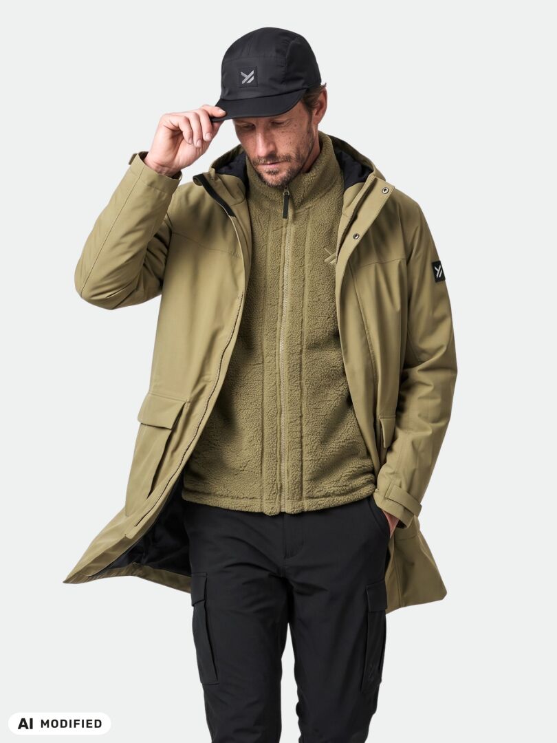 En man klädd i en Halti Kortteli DX-isolerad parka, en fleecetröja med dragkedja, svarta byxor och en keps, som står framför en enfärgad bakgrund.