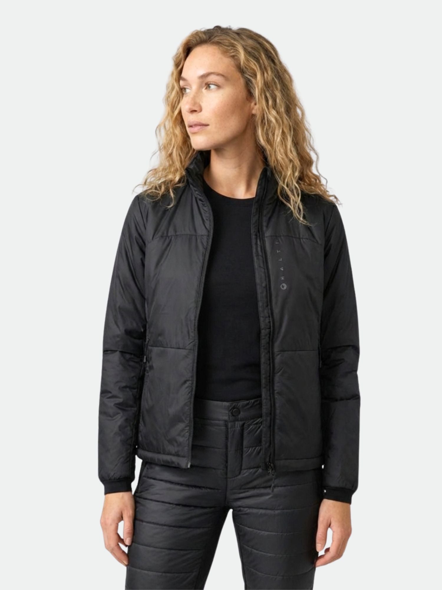 En kvinna bär Halti Neon Jacka Dam, en svart quiltad jacka med PrimaLoft-isolering, mot en enfärgad bakgrund.