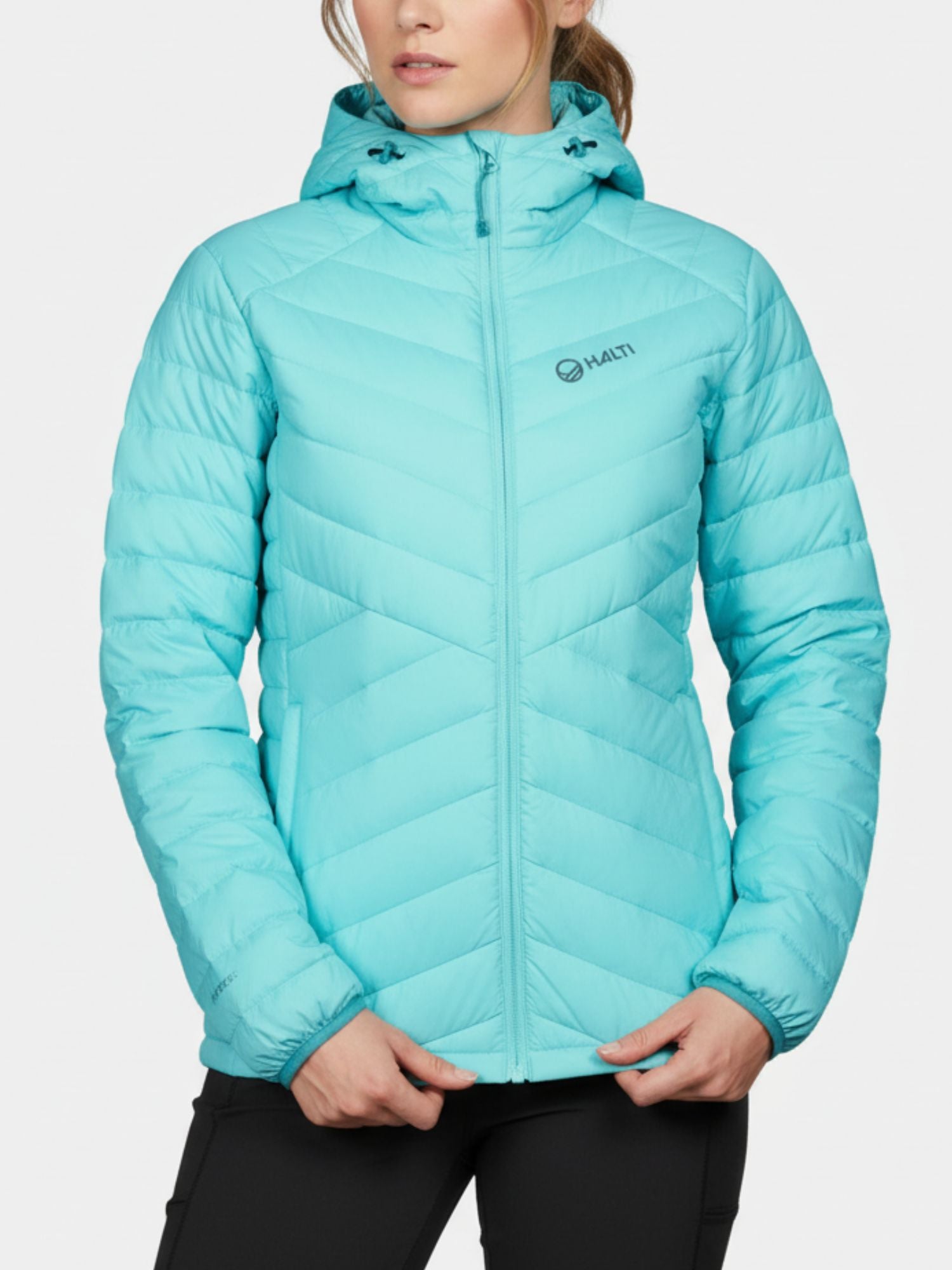 En person i en ljusblå Halti Huippu Dunjacka Dam med Pertex Quantum och svarta byxor står mot en enfärgad bakgrund.