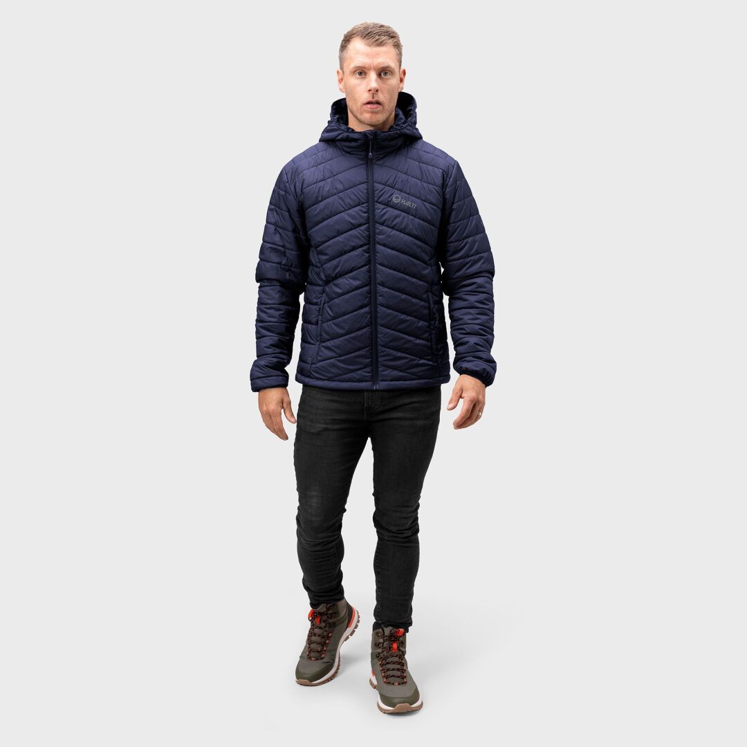 En man i Halti Komu Lätt Vadderad Jacka Herr, svarta jeans och grå vandringskängor står mot en ljusgrå bakgrund.
