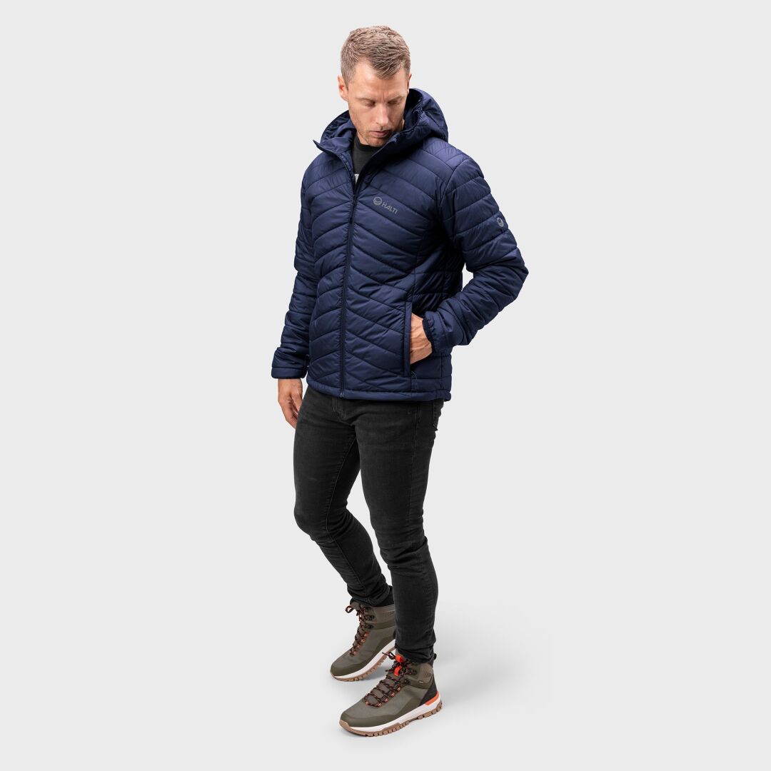 En man i en Halti Komu Lätt Vadderad Jacka Herr, svarta jeans och olivgröna vandringskängor står vid en enkel bakgrund.