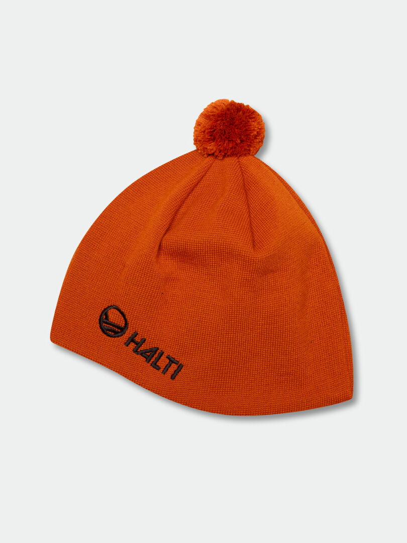 Halti Peesi Mössa är en orange stickad mössa med pom-pom och svart logotyp, designad för längdskidåkning.