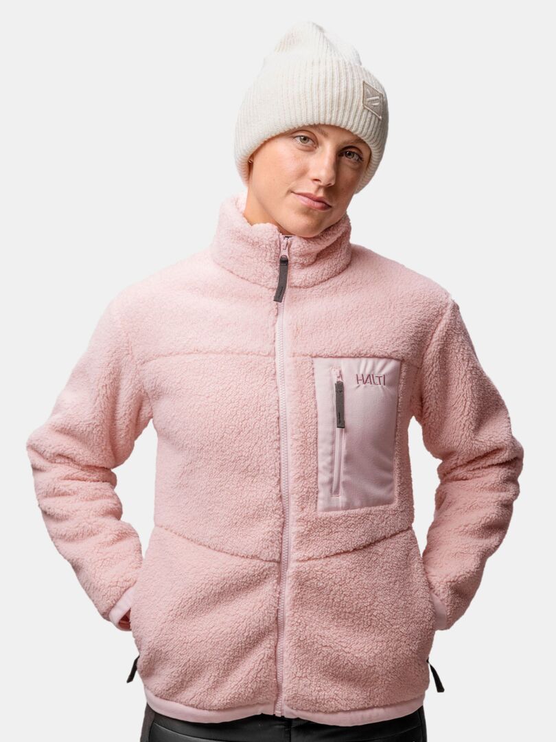 Personen har på sig en ljusrosa fleecejacka och en vit Halti Huurre Mössa, händerna i jackfickorna.