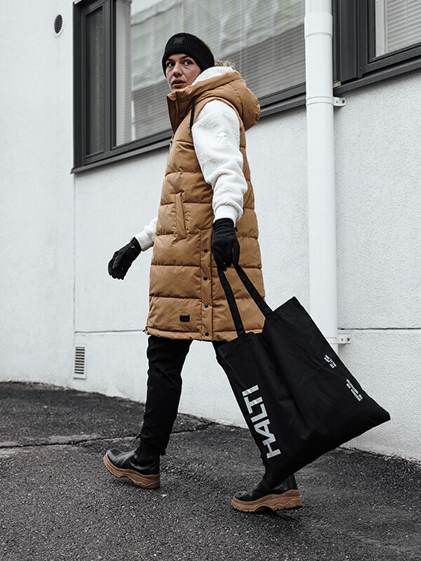 En person klädd i en lång, beige dunväst och svarta Viiri Fleece Handskar går med en svart tygkasse i handen.