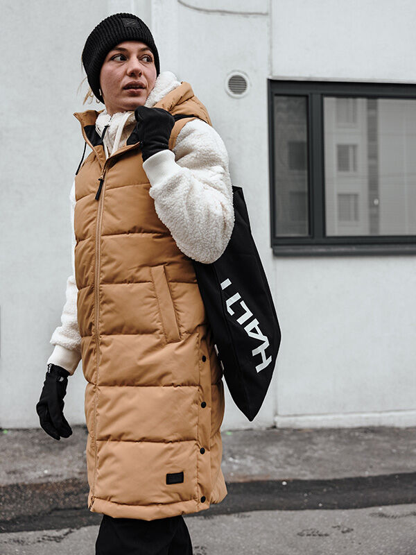 Person i brun vadderad rock och svart mössa bär Halti Viiri Fleece Handskar utanför en vit byggnad med fönster.
