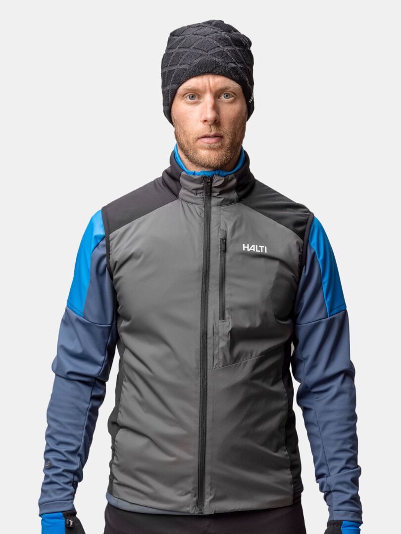 En man i grå och blå friluftsjacka och en Halti Crostie Merinomössa vänder sig mot kameran mot en enfärgad bakgrund.