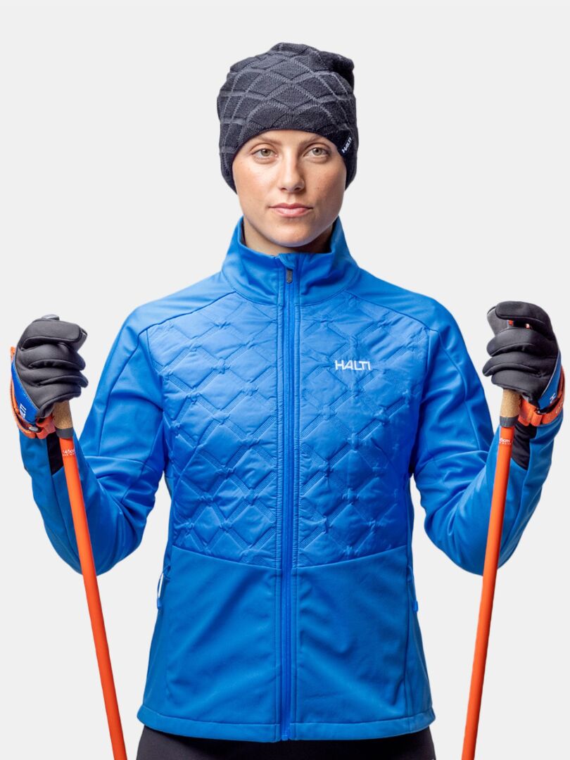 En person i blå quiltad jacka och Halti Crostie Merinomössa med orangea skidstavar, vänd framåt på vitt.