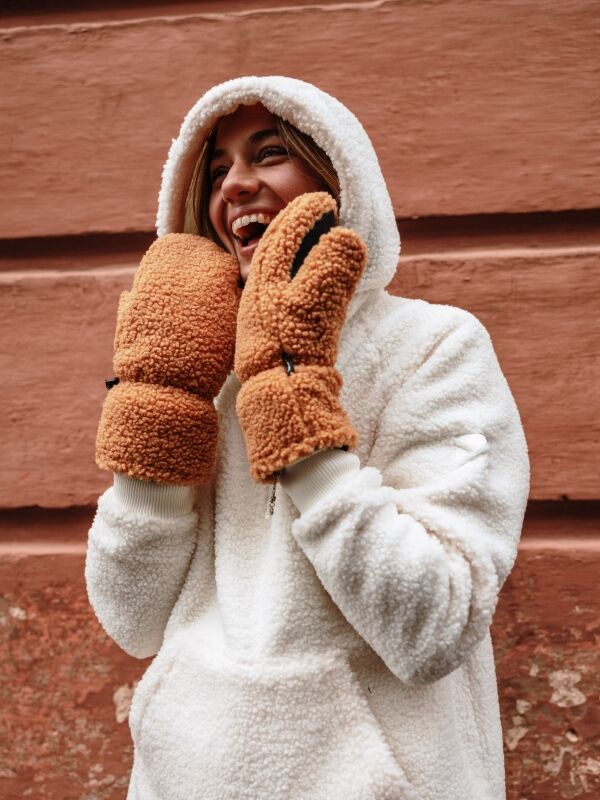 Person i fluffig vit huvtröja och Halti Lykky Fleecevantar ler framför en strukturerad brun vägg.