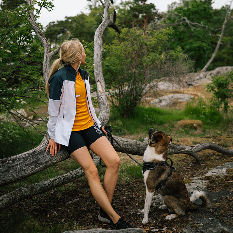 En kvinna i en Halti Vassi T-skjorta Dam sitter på ett fallet träd utomhus och håller sitt hundkoppel bredvid sig.