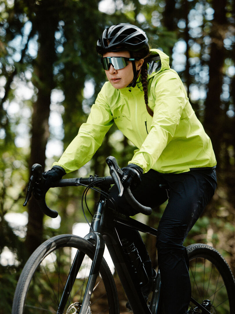 En person iförd gul jacka, hjälm och solglasögon cyklar på en svart cykel genom ett skogsområde.