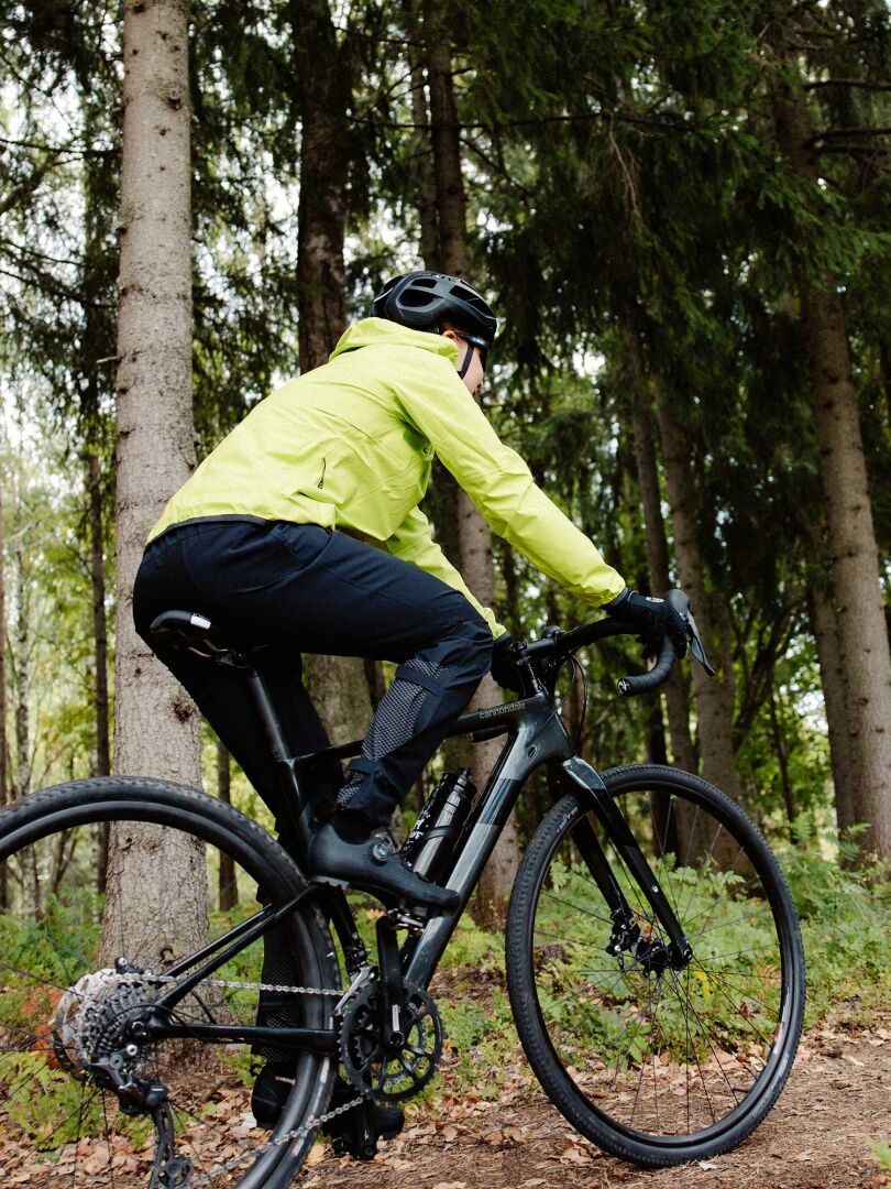 En person i en gul Halti Liike Dam 3L DrymaxX Sport Jacka cyklar på en grusväg genom en skog.