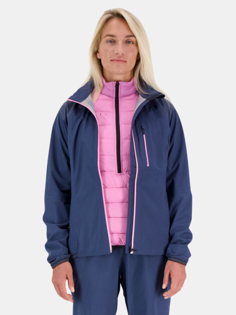 Person som står mot en slät bakgrund i en Halti Liike Dam 3L DrymaxX Sport Jacka över en rosa vadderad jacka och blå byxor.