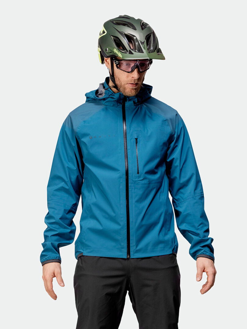 En person i en blå Halti Liike Herr 3L DrymaxX Sportjacka, svarta byxor, hjälm och solglasögon står mot en enfärgad bakgrund.