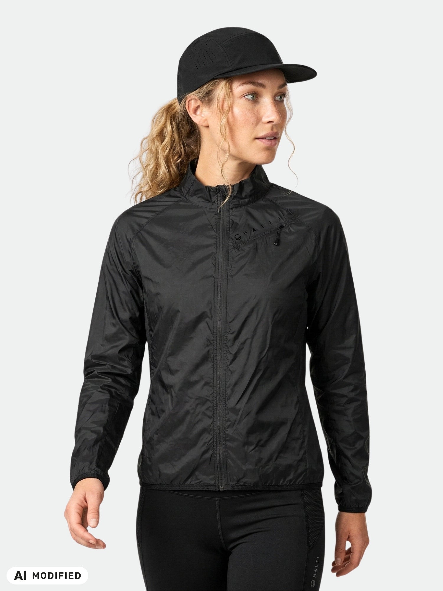 En kvinna i en Halti Control Windbreaker Jacka Dam, svart keps och leggings, som står framför en enfärgad ljus bakgrund.