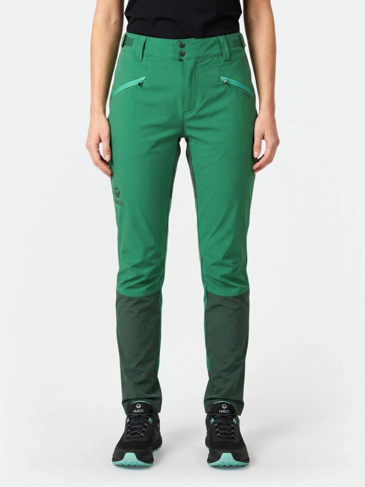 En person i Halti Tracks X-stretch Byxor Dam med mörkgröna knän och svarta skor, perfekt för utomhusträning, står ensam.