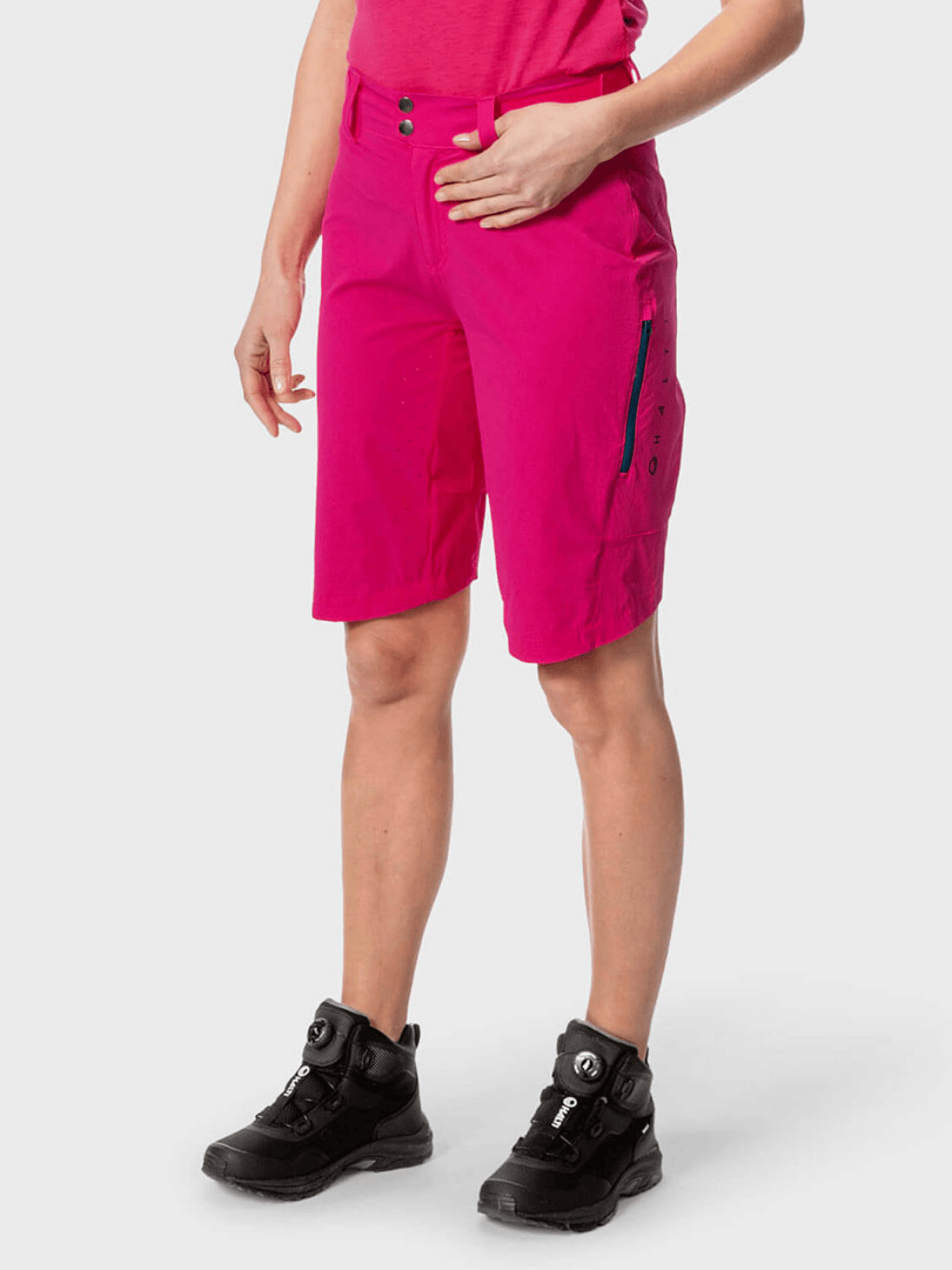 Person i Halti Tracks Dam X-stretchiga knallrosa shorts, en rosa topp och svarta vandringsskor mot en enfärgad bakgrund.