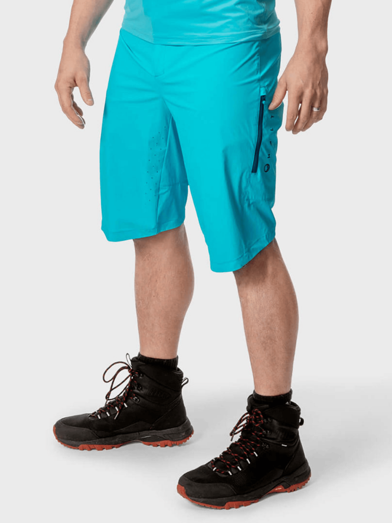 En person bär Halti Tracks Herr X-stretch shorts i klarblå och svarta vandringskängor mot en enfärgad ljus bakgrund.