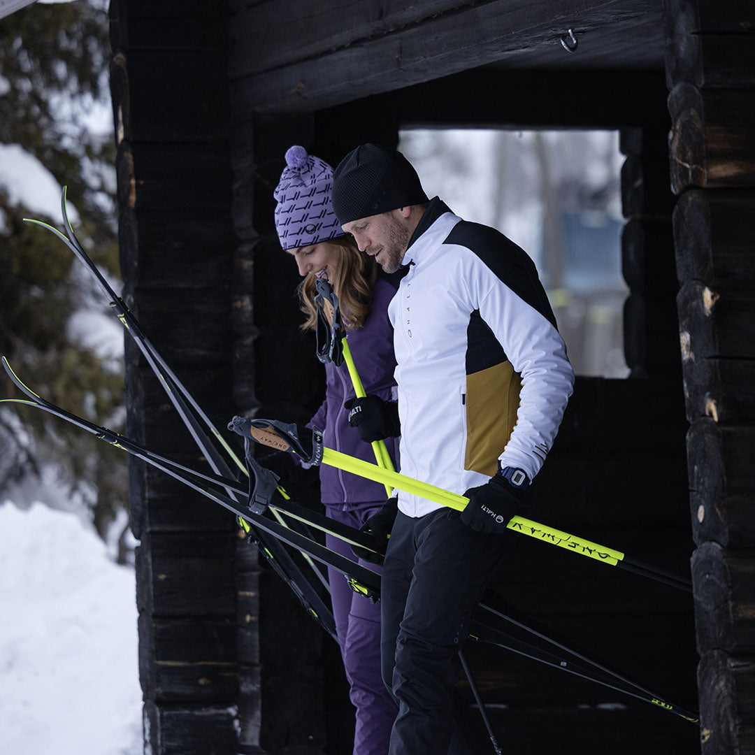 Två personer i Halti Veloce Längdskidjacka Herr håller skidor och stavar utanför en snöig träbyggnad.