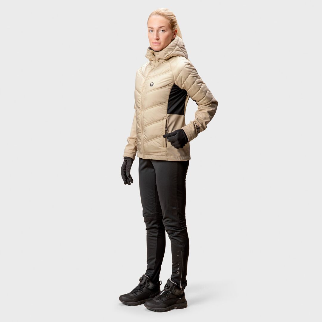 En person bär den bruna Halti Hanki Längdskidjacka Dam med svarta handskar, byxor och stövlar mot en vit bakgrund.