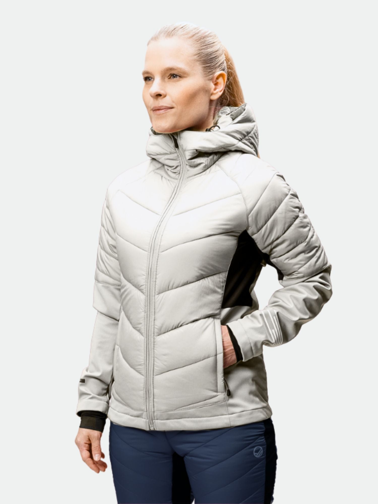 En person i vindtätt, ljusgrå Halti Hanki Längdskidjacka Dam och mörka byxor står med ena handen i fickan.