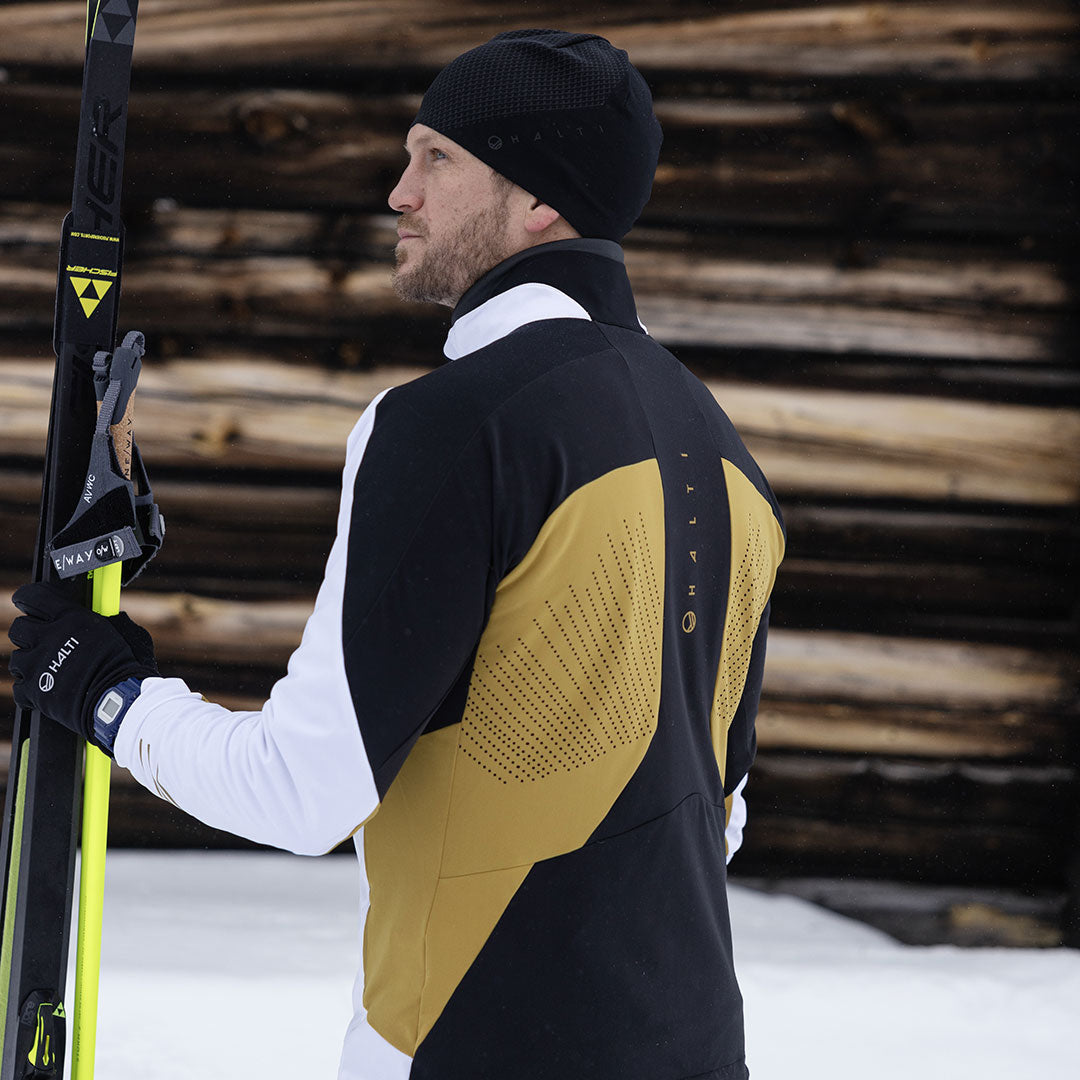 En person i Halti Veloce Längdskidjacka Herr håller skidor, stående på snö framför staplade stockar, vänd bort.