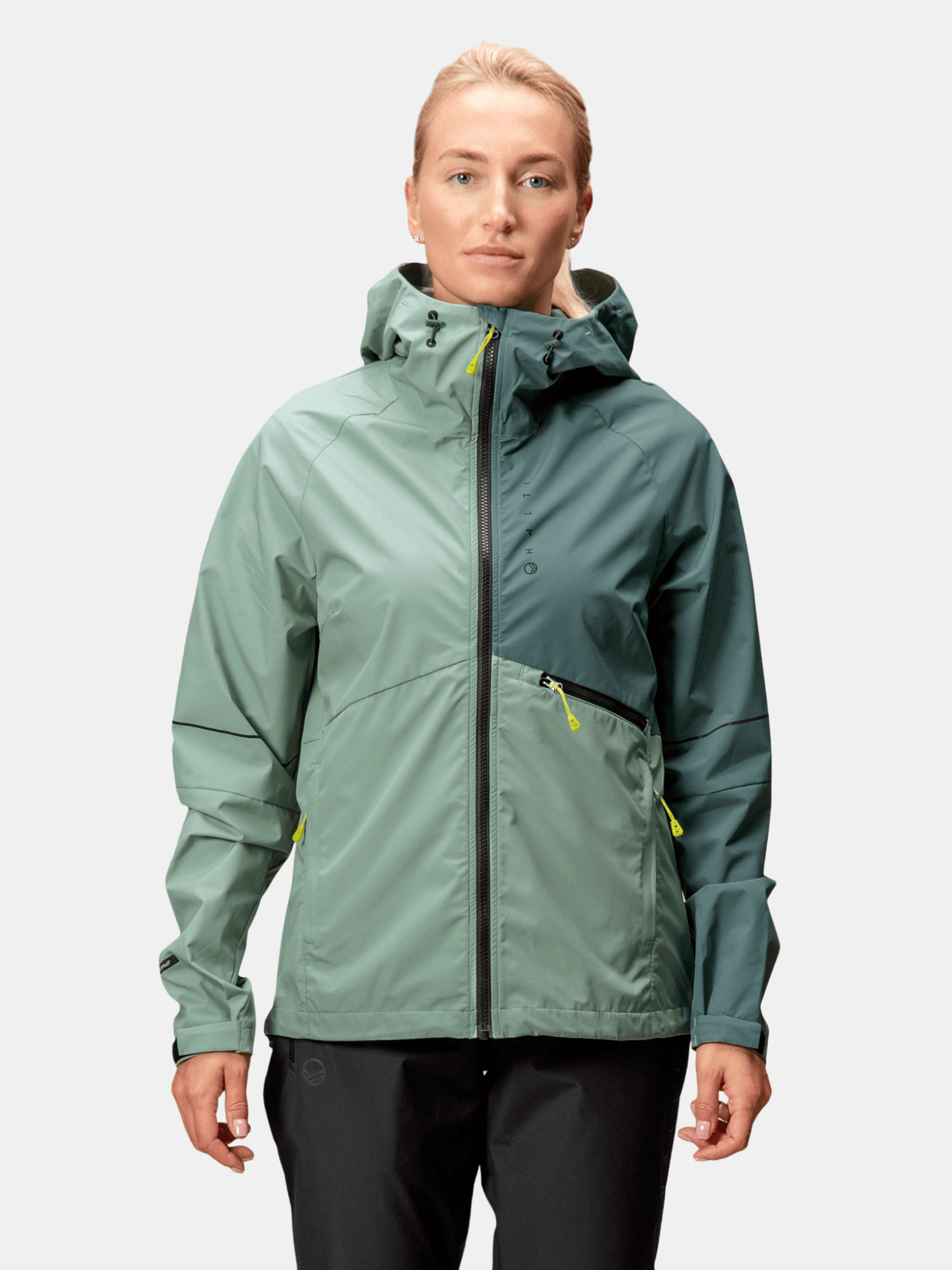 En kvinna bär Halti Shelter Stormwall 3L Jacka Dam i grönt, med fickor med dragkedja, mot en enfärgad bakgrund.