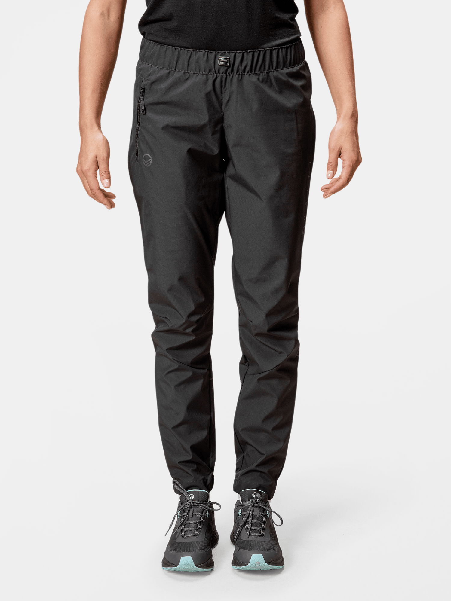 En person bär svarta Halti Shelter Stormwall 3L Byxor Dam-byxor, en svart skjorta och blågrönt färgade sneakers mot en enfärgad bakgrund.