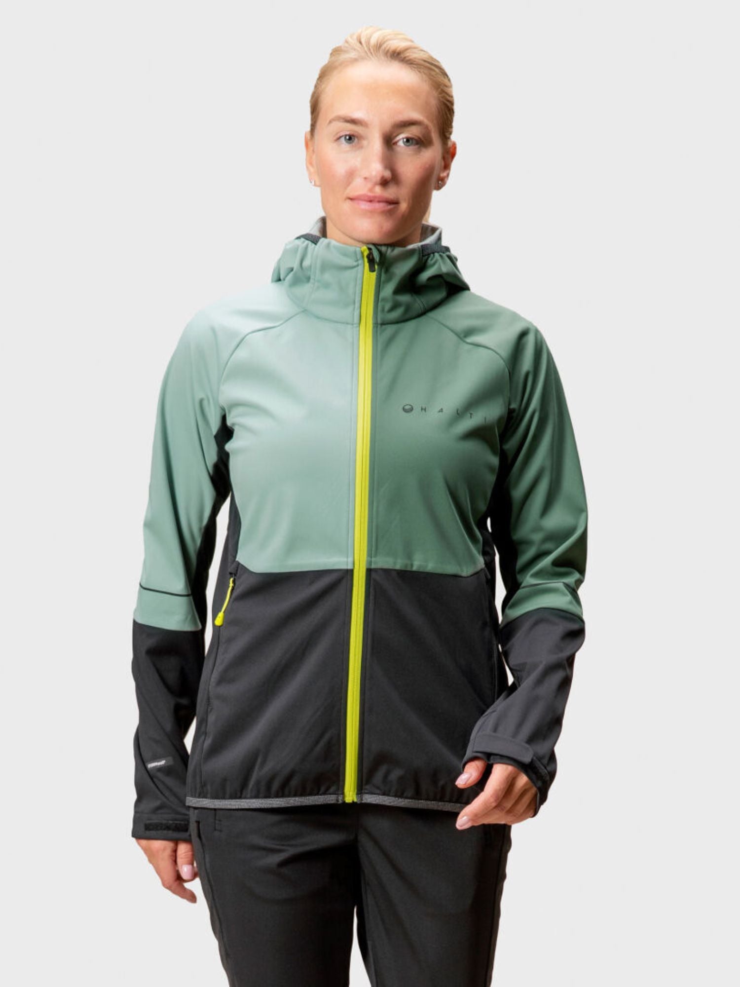 En person bär Halti Exhale Stormwall Jacka Dam i grönt och svart med gult blixtlås mot en ljus bakgrund.
