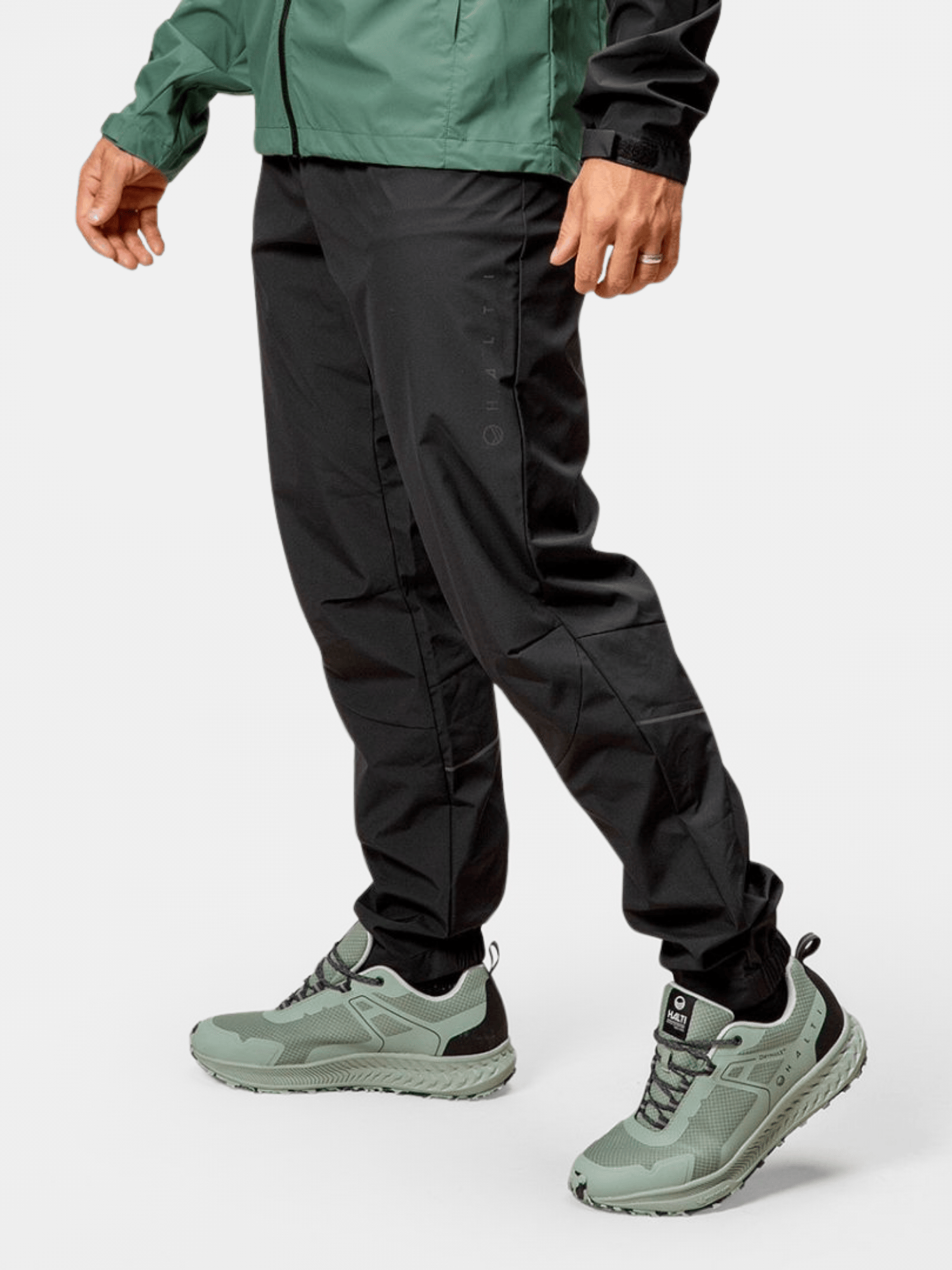 En person i Halti Shelter Stormwall 3L Byxor Herr, grön jacka och ljusgröna sneakers står mot en enfärgad bakgrund.